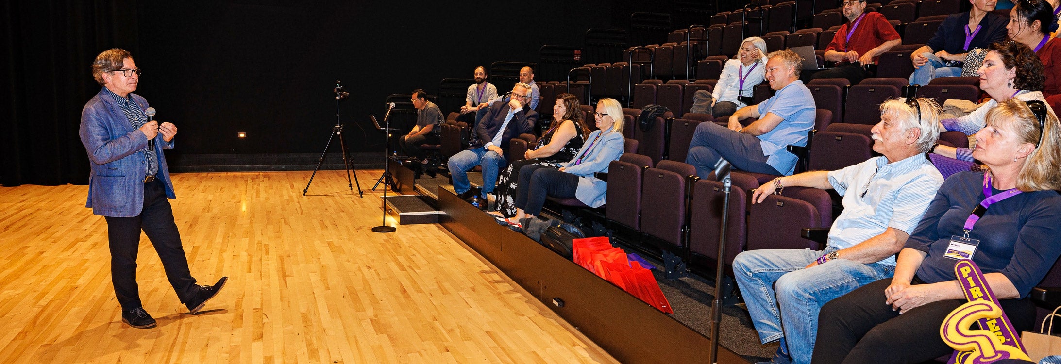 A man in a gray jacket speaking from a stage inside the Black Box theater.