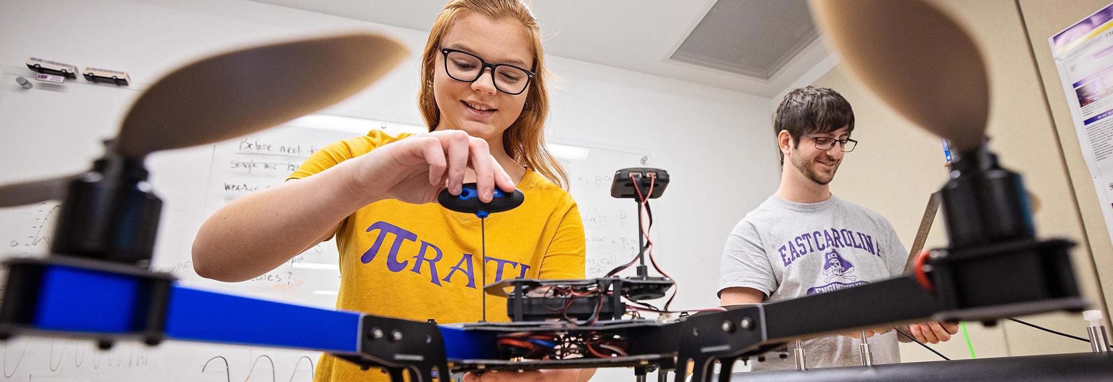 A student in a a gold t-shirt works on a drone while another student looks on.